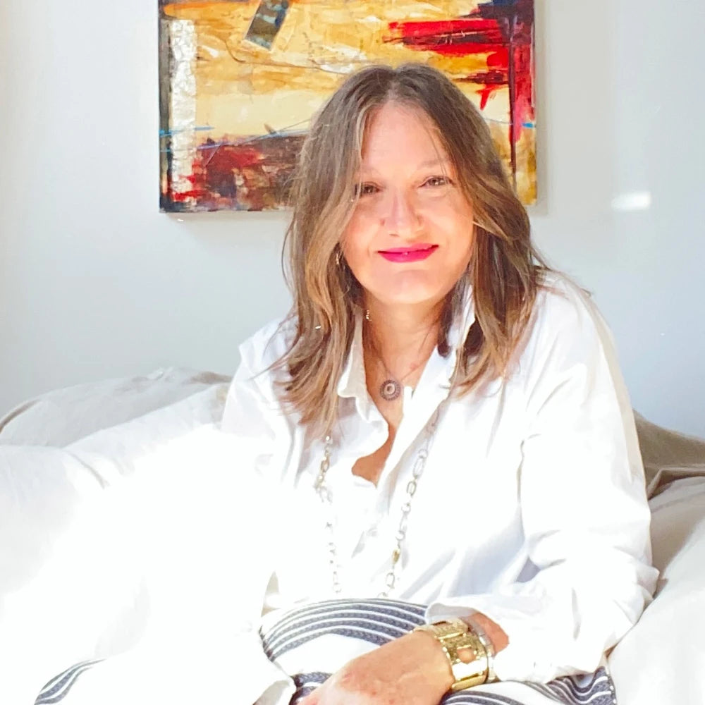 Manuela Valenti founder of By Valenti in a white shirt sitting on a couch with one of her own colorful abstract painting in the background
