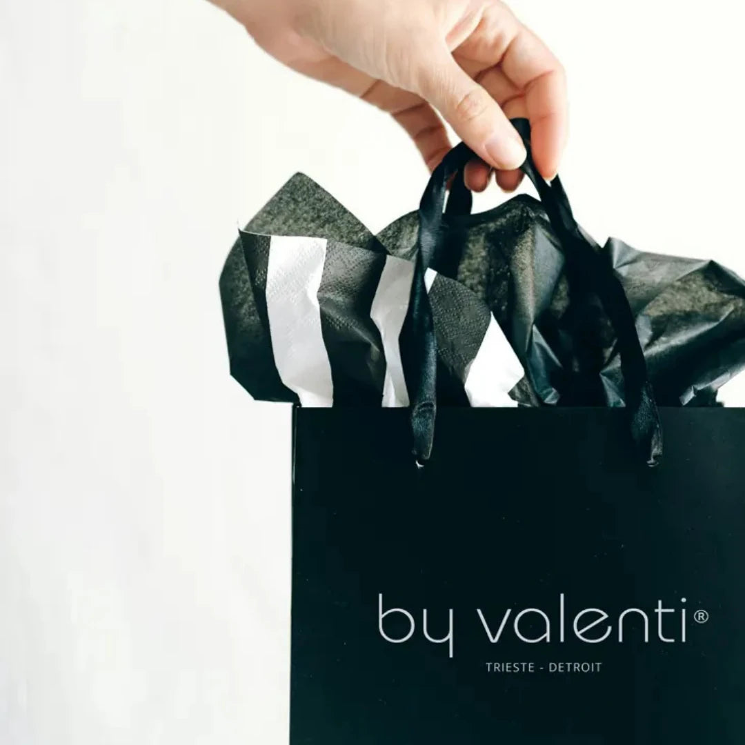 Hand holding a black 'by valenti' shopping bag with ribbons against a white background