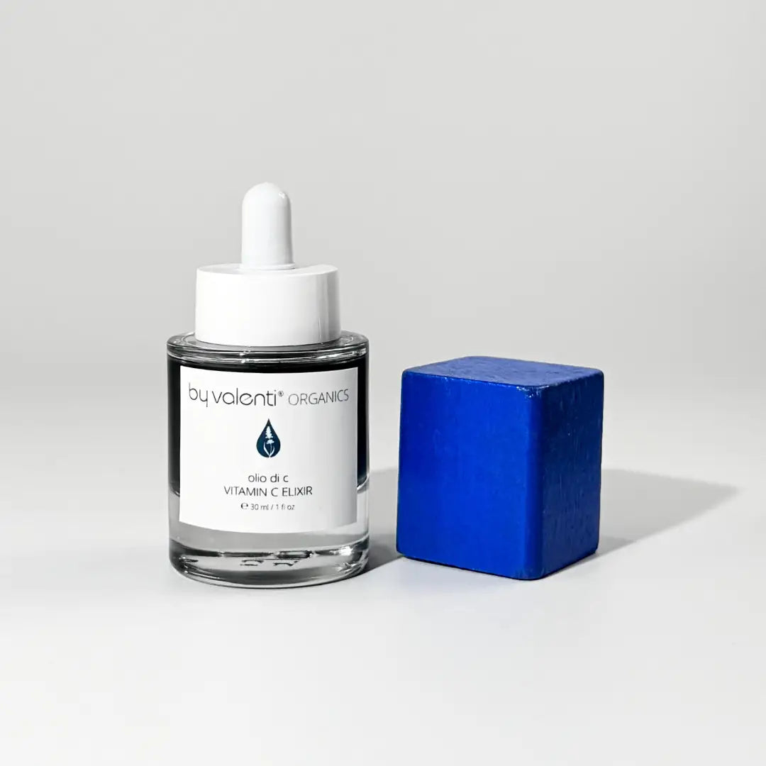 Glass dropper bottle of BY VALENTI blue vitamin C elixir next to a blue cube on a white background