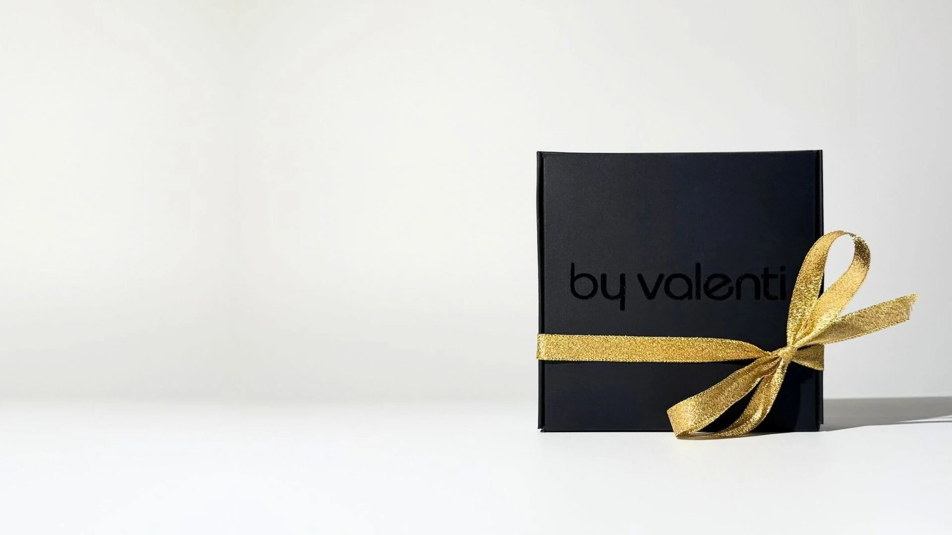 Black gift box with gold ribbon and 'by valenti' branding on a white background