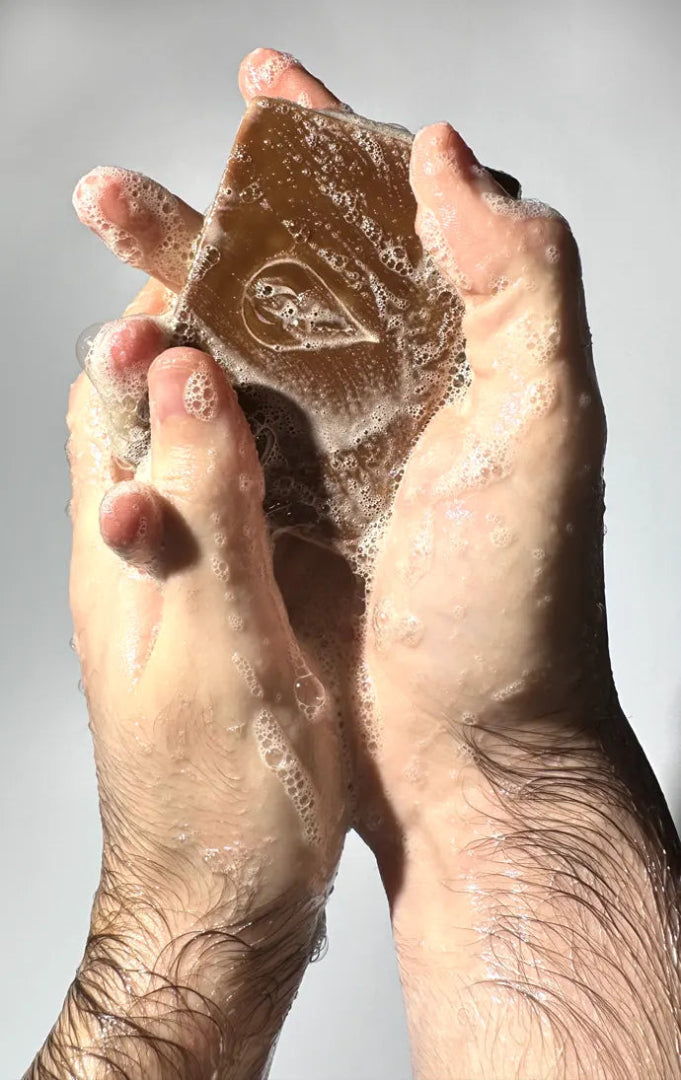 Man hands holding a BY VALENTI Dark Raw Chocolate Bar Soap with lather and visible logo in front of a light gray background
