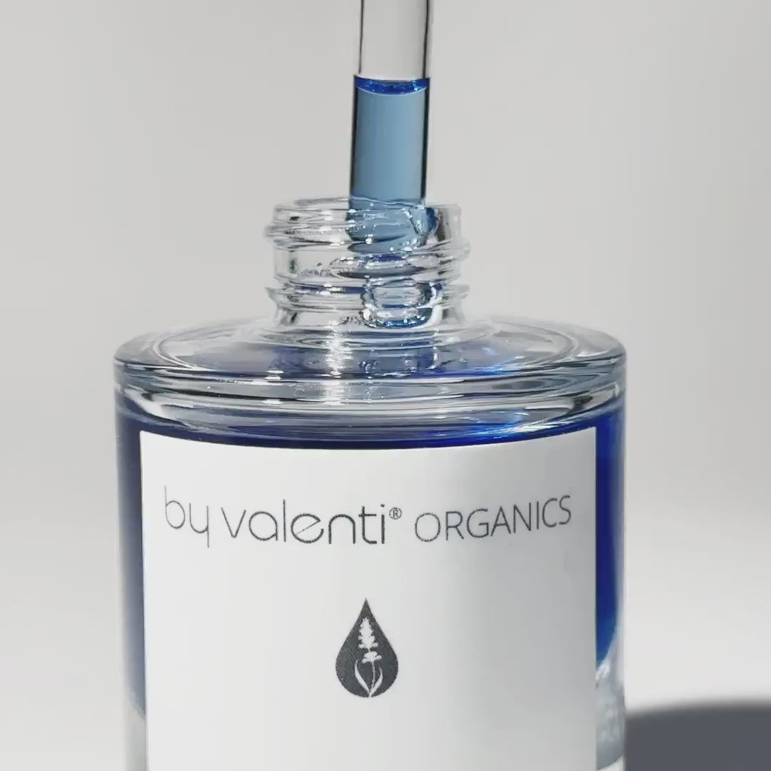 A dropper dripping By Valenti Organics Olio di C blue colored Vitamin C Serum into the bottle on a white background