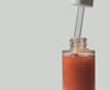 By Valenti red-orange shimmering body Oil dripping from a dropper into the bottle on a white background